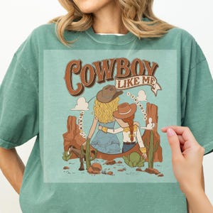 Pu&ograve; includere: Una t-shirt verde salvia con un'illustrazione grafica di due figure con cappelli da cowboy in un paesaggio desertico con formazioni rocciose rosse, un cavallo e cactus. Il testo "COWBOY LIKE ME" e "I KNEW YOU" &egrave; stampato in marrone.
