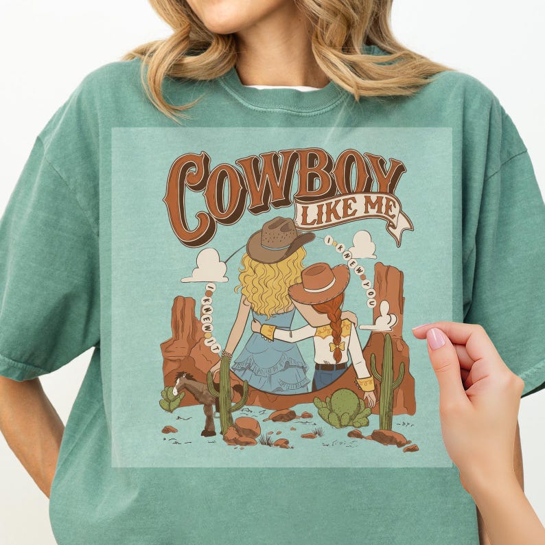 Pu&ograve; includere: Una t-shirt verde salvia con un'illustrazione grafica di due figure con cappelli da cowboy in un paesaggio desertico con formazioni rocciose rosse, un cavallo e cactus. Il testo "COWBOY LIKE ME" e "I KNEW YOU" &egrave; stampato in marrone.