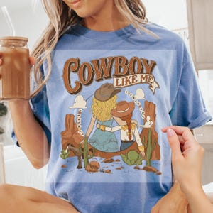 Pu&ograve; includere: Una t-shirt azzurra a girocollo con una stampa grafica di due figure con cappelli da cowboy in un paesaggio desertico con cactus, un cavallo e il testo "COWBOY LIKE ME" e "I KNEW YOU" scritto in un banner curvo e in un motivo circolare.