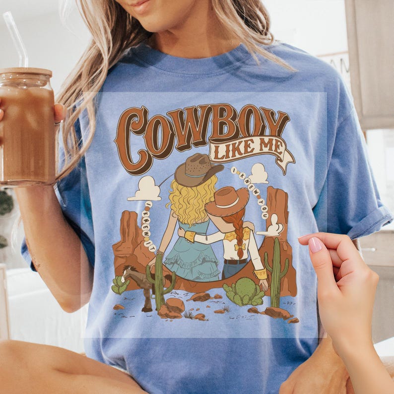 Pu&ograve; includere: Una t-shirt azzurra a girocollo con una stampa grafica di due figure con cappelli da cowboy in un paesaggio desertico con cactus, un cavallo e il testo "COWBOY LIKE ME" e "I KNEW YOU" scritto in un banner curvo e in un motivo circolare.