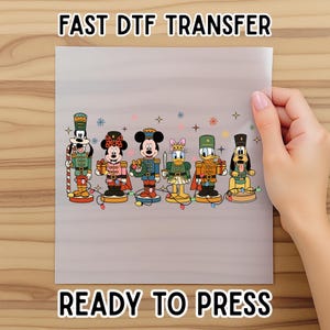May include: A gray transfer sheet featuring cartoon characters dressed as nutcrackers. The characters include Mickey Mouse, Minnie Mouse, Donald Duck, Daisy Duck, Goofy, and Pluto. The text "FAST DTF TRANSFER" and "READY TO PRESS" are displayed.