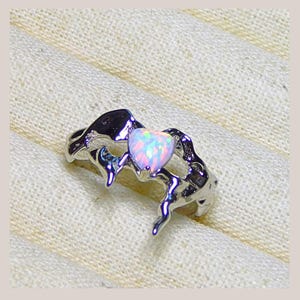 May include: A silver ring with an opal heart-shaped stone. The ring has a unique, flowing design with a textured appearance. The opal stone displays a range of colors, including pink, blue, and white, creating a shimmering effect.