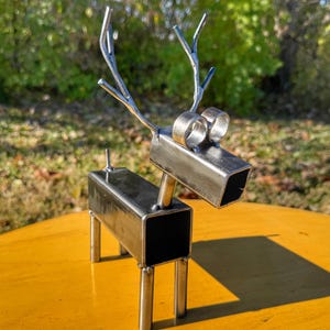 May include: A metal sculpture of a deer with a rectangular body and head, and cylindrical eyes. The antlers are made of curved metal rods. The sculpture is silver and sits on a yellow surface.