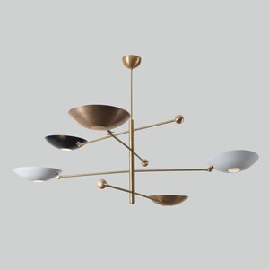 May include: Modern brass chandelier with five bowl-shaped light fixtures. The bowls are in various colors, including gold, black, and white. The fixture has a central vertical rod with horizontal arms extending outwards, each holding a light.