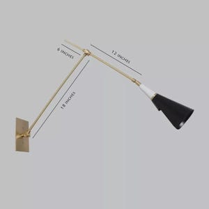 May include: A wall-mounted, adjustable lamp with a brass finish. The lamp features a black cone-shaped shade with a white accent. The arm extends 18 inches, with a 6-inch and 12-inch extension. The lamp is mounted on a rectangular brass plate.