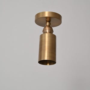 May include: A cylindrical, brass-colored ceiling light fixture. The fixture has a circular base and a cylindrical shade. The light is suspended from the ceiling by a short, decorative stem. The overall design is minimalist and modern.