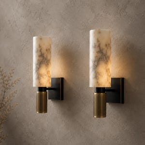 May include: Two wall sconces featuring cylindrical white marble shades with grey veining. Each light has a matte black square wall mount and a vertical, ribbed brass-toned metal accent piece connecting the shade to the fixture base.