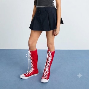 Puede incluir: Una camiseta sin mangas de color gris oscuro con una fina raya horizontal plateada en la cintura, combinada con una minifalda plisada negra lisa y botas altas de lona roja con cordones blancos, punteras y suelas de goma blanca.