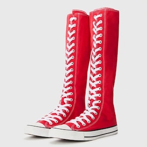 May include: A pair of knee-high red canvas trainers featuring a tall shaft, white laces running up the entire front, white rubber toe caps, and white rubber soles with a thin black stripe detail along the edge of the base of the footwear.