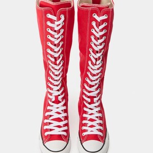 Puede incluir: Un par de botas altas de lona roja con cordones blancos que se extienden desde la punta hasta la parte superior de la ca&ntilde;a. Las botas tienen una puntera de goma blanca, una suela de goma blanca con una fina raya negra y una cremallera vertical en la parte posterior.