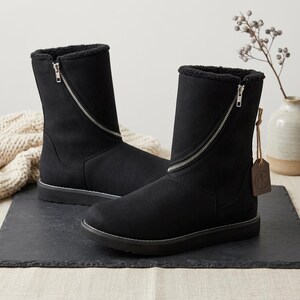 Könnte beinhalten: Ein Paar schwarze halbhohe Stiefel mit einem diagonalen silbernen Reißverschluss, der vom oberen Schaft bis zum Spann verläuft. Die Stiefel haben ein schwarzes Innenfutter aus Kunstfell und eine flache schwarze Sohle mit weißen Ziernähten.