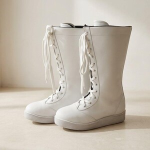 Puede incluir: Un par de botas de media caña en color blanco sólido con acabado liso. Estas botas cuentan con un cierre frontal de cordones blancos a juego, puntera redondeada y una suela plana de color claro, con un diseño de caña alta y líneas limpias.