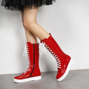 May include: A pair of mid-calf boots in glossy red patent material featuring white laces that run up the front, a black side zipper, and a thick white rubber sole. The boots have a rounded toe and a structured shaft that reaches below the knee.
