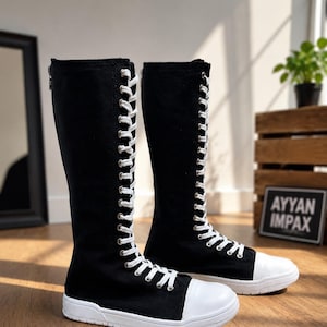 Puede incluir: Un par de botas altas de lona negra con punteras de goma blanca, suelas de goma blanca y cordones blancos que suben verticalmente por la parte delantera. Las botas incluyen ojales de metal plateado y un cierre de cremallera en la parte trasera.