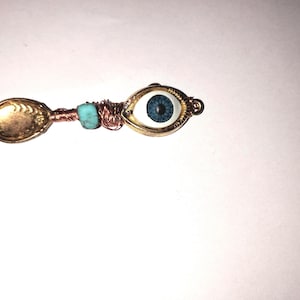 Puede incluir: Un colgante artesanal en forma de cuchara con un cuenco color latón y un dije decorativo de ojo. El dije de ojo tiene un iris azul y un marco dorado. El mango está envuelto con alambre de cobre y adornado con cuentas turquesas.