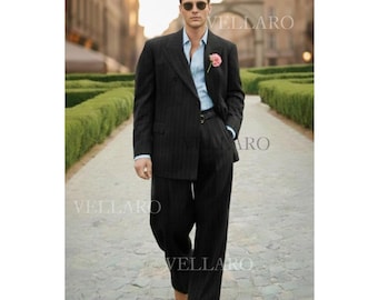 May include: A dark gray pinstripe suit with a light blue shirt and a pink flower. The suit has a double-breasted jacket and wide-leg trousers. The person is wearing sunglasses and walking on a cobblestone path.