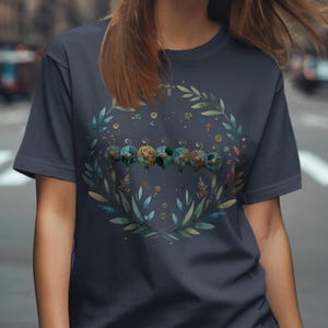 以下が含まれることがあります： ネイビーブルーのTシャツ。水彩画のデザインで、葉と小さなカラフルなオーブのリースが描かれています。デザインはシャツの中央に配置され、背景はぼやけています。シャツは柔らかい素材で作られています。