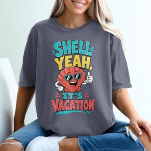 May include: A charcoal gray t-shirt featuring a graphic of a smiling, anthropomorphic red seashell wearing teal sunglasses and giving a thumbs up. The text reads "SHELL YEAH, IT'S VACATION" in bold, colorful lettering with teal, yellow, and red accents.