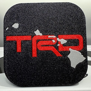 May include: A black square car emblem with a red "TRD" logo and a white outline of the Hawaiian islands.