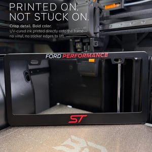 Peut inclure: Un cadre de plaque d'immatriculation noir avec le texte blanc FORD PERFORMANCE en haut et un logo ST rouge en bas. Le cadre est placé devant une imprimante 3D, avec le texte au-dessus indiquant NEW UV-PRINTED GRAPHICS PRINTED ON. NOT STUCK ON.