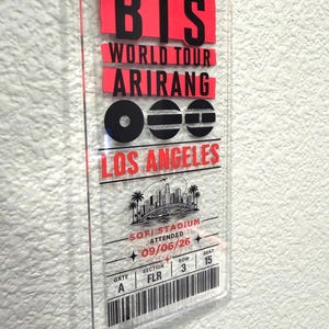 May include: A rectangular clear acrylic souvenir concert ticket for a BTS World Tour Arirang event in Los Angeles at SoFi Stadium. The ticket features black and red text, a city skyline graphic, and details including date 09/06/26, Gate A, Section FLR, Row 3, Seat 15.