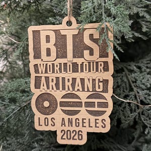 May include: A rectangular wooden ornament with a natural finish, featuring laser-engraved text that reads BTS WORLD TOUR ARIRANG LOS ANGELES 2026, accompanied by three circular icons with horizontal line details, suspended by a twine loop.