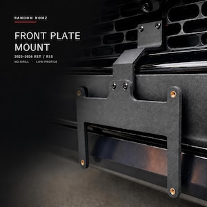 May include: A black matte front license plate mount for 2022-2026 R1T and R1S vehicles. The bracket features a low-profile, no-drill design with visible mounting screws and four brass-colored threaded inserts for secure license plate attachment.