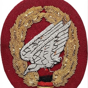 Op de afbeelding: Een ovaalvormige geborduurde patch met een donkerrode vilten achtergrond. In het midden bevindt zich een zilverkleurig vleugelembleem omringd door een gedetailleerde goudkleurige lauwerkrans. Aan de basis van de krans zitten kleine horizontale banden van zwart, rood en goudkleurig garen.