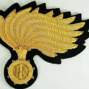 May include: A gold-colored embroidered patch with a black felt backing. The patch features a stylized design of a flame or feather shape, with a circular emblem at the base. The emblem has a detailed design with the letters "R" and "H".