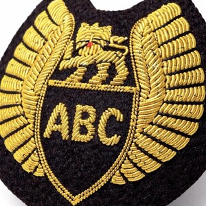 Op de afbeelding: Een zwart en goud geborduurd embleem met de letters "ABC" in het midden. Het ontwerp omvat een schild, gouden vleugels en een leeuwenembleem. Dit is waarschijnlijk een militair of luchtvaartinsigne.