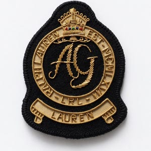 Peut inclure: Écusson brodé noir avec lettrage et détails dorés. Le patch présente les initiales "AG" dans une écriture stylisée à l'intérieur d'un cercle doré, entouré de texte. Une couronne dorée est placée au-dessus du cercle. Le patch a une bordure noire.