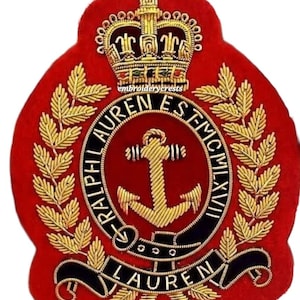 Könnte beinhalten: Ein rotes, gesticktes Wappen mit goldenen Details, das eine Krone, Lorbeerblätter und einen Anker zeigt. Der Text "RALPH LAUREN EST MCMLXVII" ist in Gold und Schwarz gestickt. Das Wappen hat ein geschwungenes schwarzes Banner mit dem Namen "LAUREN".