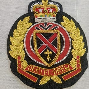 May include: A black embroidered patch featuring a gold crown at the top, gold laurel leaves on the sides, and a central shield with a red cross. A red banner at the bottom displays the gold text OERTEL-DREW in a bold, capitalized font.