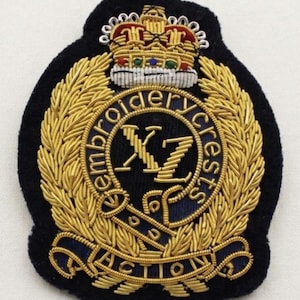 May include: A black embroidered patch with gold-colored thread detailing. The patch features a crown, the text "Embroidery Crests", and the word "Action". The design includes a circular emblem with the letters "XZ".