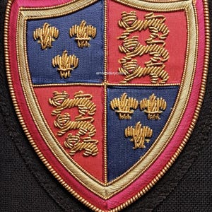 May include: Embroidered shield patch with a heraldic design. The shield is divided into four quadrants: two blue and two red. Gold thread outlines the shield and embellishes the symbols within, including stylized lions and floral motifs. The patch has a pink border.