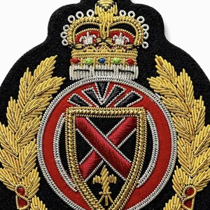 May include: A black felt embroidered patch featuring a gold crown with colorful accents at the top. Below the crown is a red circular emblem containing a shield with a red cross, flanked by gold laurel branches made of metallic thread.