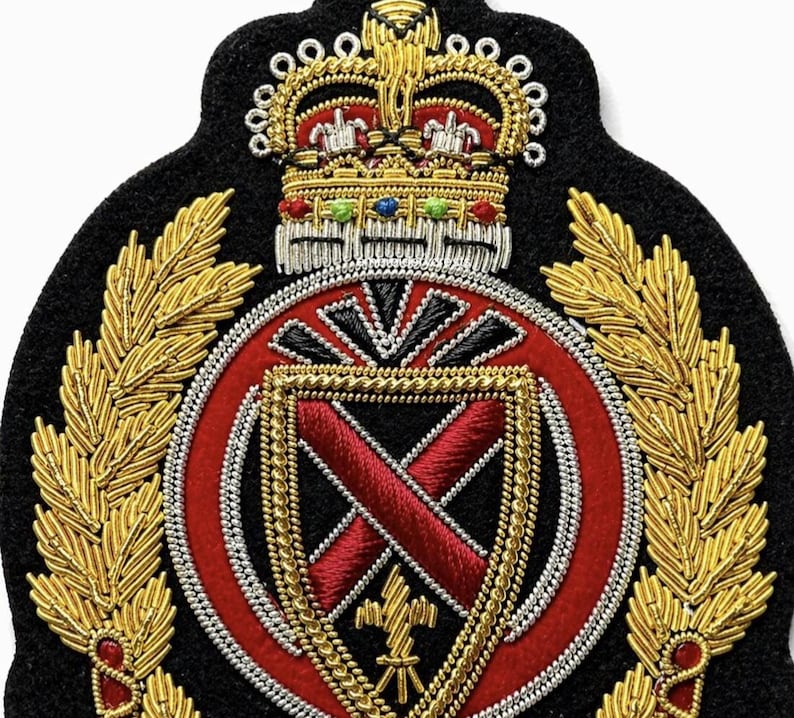 May include: A black felt embroidered patch featuring a gold crown with colorful accents at the top. Below the crown is a red circular emblem containing a shield with a red cross, flanked by gold laurel branches made of metallic thread.