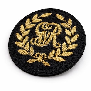 May include: A round, black embroidered patch featuring a gold-colored monogram within a laurel wreath. The monogram appears to be two intertwined letters. The patch has a textured appearance and is set against a white background.