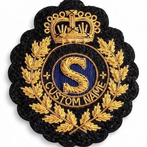 Op de afbeelding: Een zwart geborduurd embleem met gouden details. Het embleem heeft een gouden kroon, een blauwe cirkel met een gouden letter "S" en de woorden "CUSTOM NAME". Gouden bladeren omringen de cirkel.