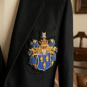 Puede incluir: Un blazer negro con un escudo bordado en el bolsillo. El escudo presenta un escudo azul con detalles dorados y amarillos, una corona y el texto "victorycrests". Se ve un botón dorado.
