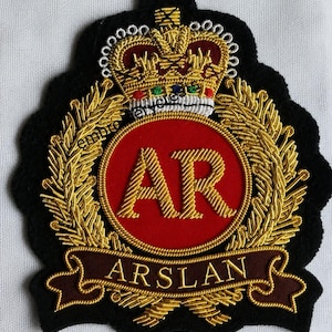 May include: A black embroidered patch featuring a gold crown with red, green, and blue accents above a red circular emblem containing the gold letters AR. Below the circle is a dark red banner with the gold text ARSLAN, all framed by gold laurel leaves.
