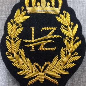 Puede incluir: Parche bordado negro con corona dorada, corona de laurel y letras estilizadas. El parche tiene una forma única y probablemente está diseñado para ropa o accesorios. El texto "embroiderycrests" es visible en la corona.