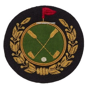 May include: A circular black embroidered patch featuring a golf-themed design. The center showcases crossed golf clubs and a golf ball within a green circle. A red flag and gold laurel wreath complete the design. The text "embroiderycrests" is visible.