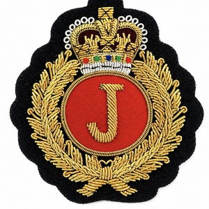 May include: A black embroidered patch featuring a central red circle with a gold letter J. The design is surrounded by a gold laurel wreath and topped with a detailed gold crown featuring small red, blue, and green accents on a black base.