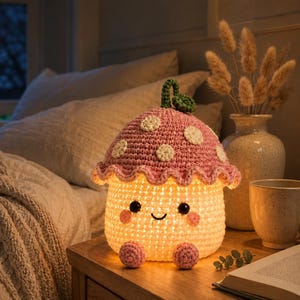 May include: A crocheted mushroom-shaped lamp with a pink cap featuring white spots and a green stem top. The base is cream-coloured with a glowing interior, black bead eyes, a stitched smile, pink cheeks, and two small pink feet at the bottom.
