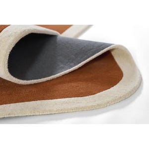 May include: Close-up of a brown and cream-colored area rug with a curved design. The rug's edge is cream-colored, framing the brown center. The underside of the rug is visible, showing a dark gray backing.