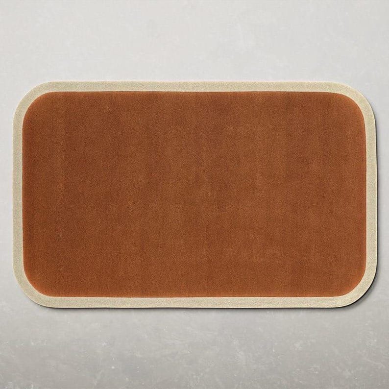 May include: A rectangular, rust-colored rug with rounded corners and a cream-colored border. The rug has a soft, textured appearance, suitable for a living room or bedroom. The rug's dimensions are not visible.