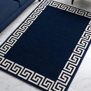 May include: A rectangular navy blue rug with a white Greek key border. The rug is placed on a light-colored floor, and a blue sofa and white chair are partially visible in the background. The rug's dimensions are not visible.