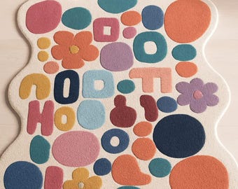 May include: A cream-colored rug with a whimsical design featuring colorful shapes and letters. The rug has a wavy outline and is adorned with circles, squares, flowers, and the letters "GOOD HOO". The shapes are in shades of pink, orange, blue, and yellow.