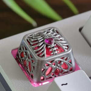 May include: A silver-colored keyboard keycap with a skeleton design. The keycap features a rib cage design with a pink heart-shaped gem in the center. The keycap sits on a pink base and is surrounded by other keyboard keys.